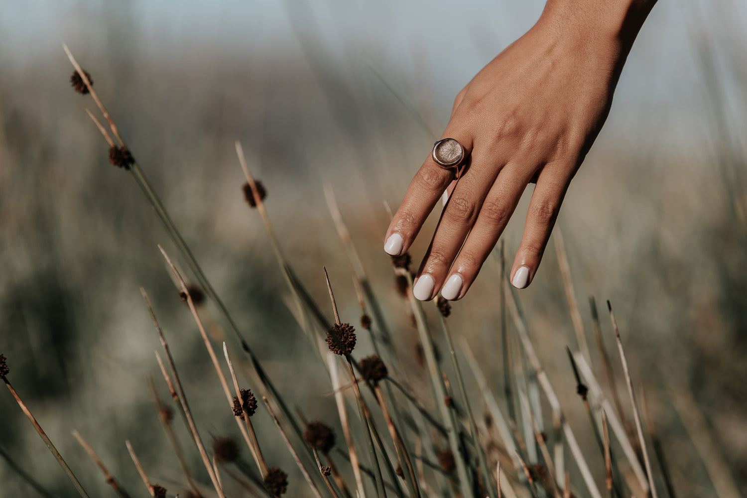 An artful photo of a lady's hand, wearing a fingerprint impression ring on her index finger, over some grasses.