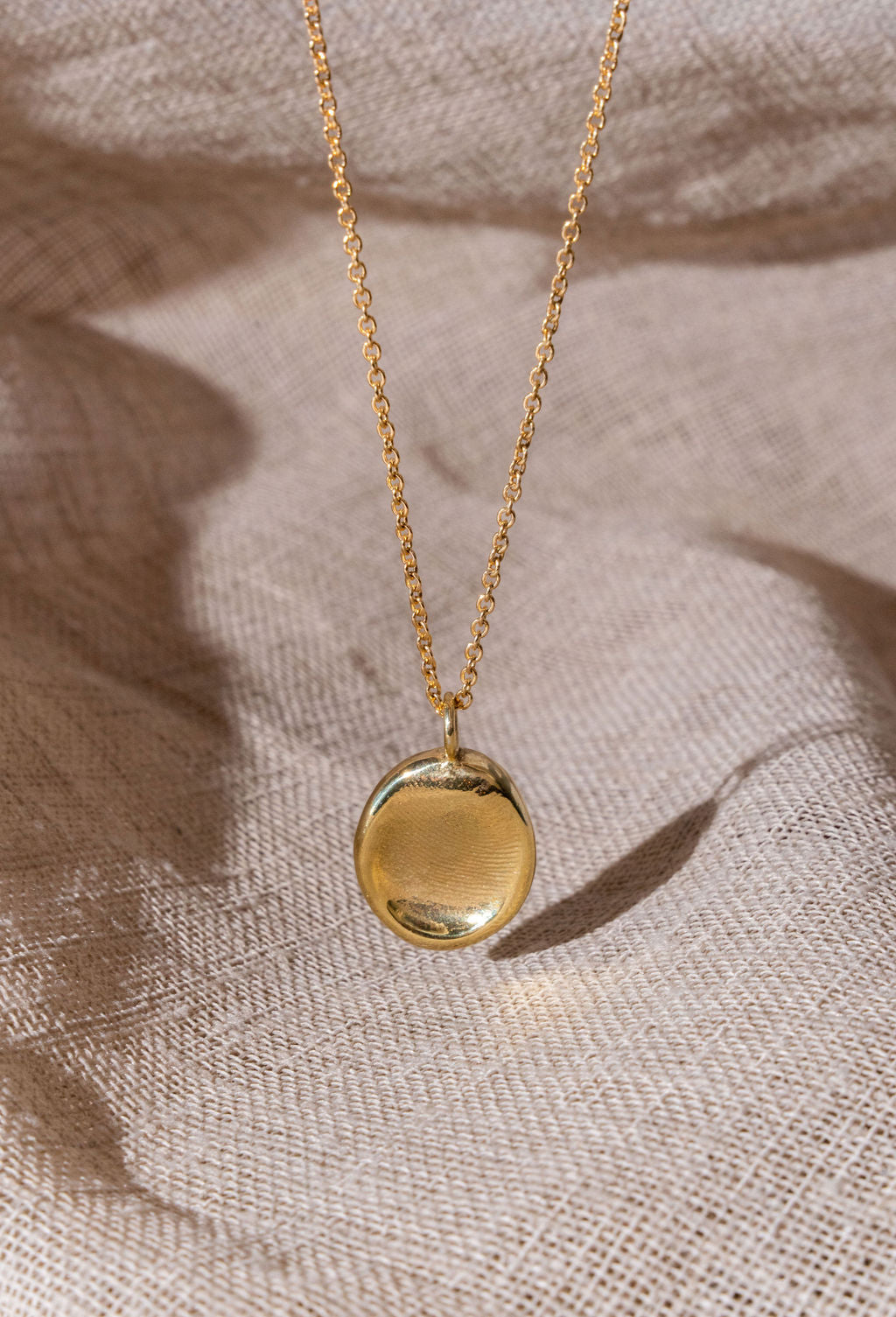 A gold fingerprint impression necklace dangling over natural linen.