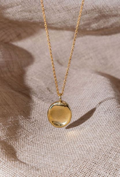 A gold fingerprint impression necklace dangling over natural linen.