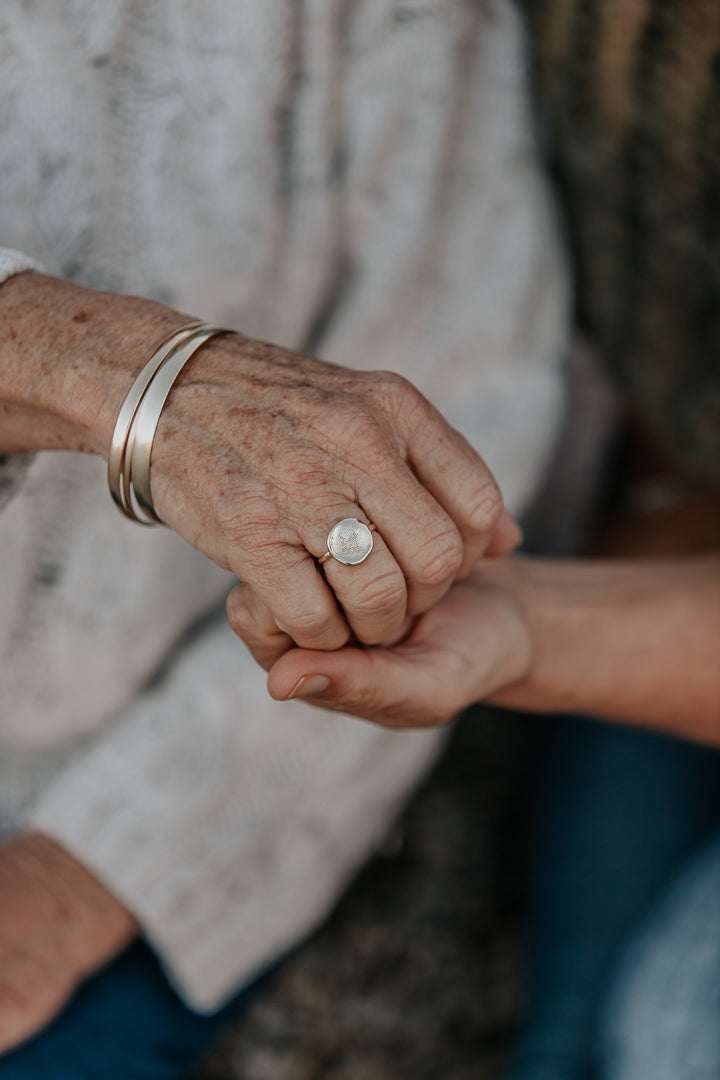 A close-up of an older mother and her daughter holding each others' hand, while the mother wears an impression ring with her daughter's fingerprint on it. 